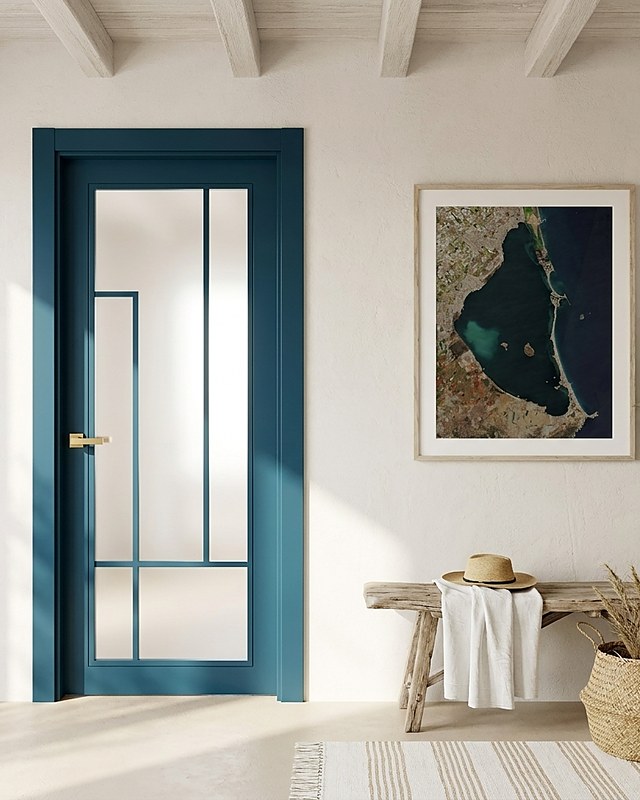 Puerta vidriera lacada en azul petróleo con montantes y cristal mate, manilla dorada, en vivienda mediterránea encalada con vigas y lámina aérea del Mar Menor. Imagen ilustrativa.