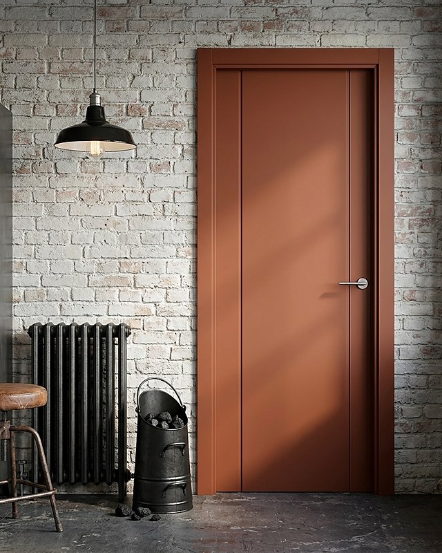 Puerta lacada en color teja con dos cuerpos verticales y manilla de acero, sobre pared de ladrillo encalado con radiador de hierro. Imagen ilustrativa.